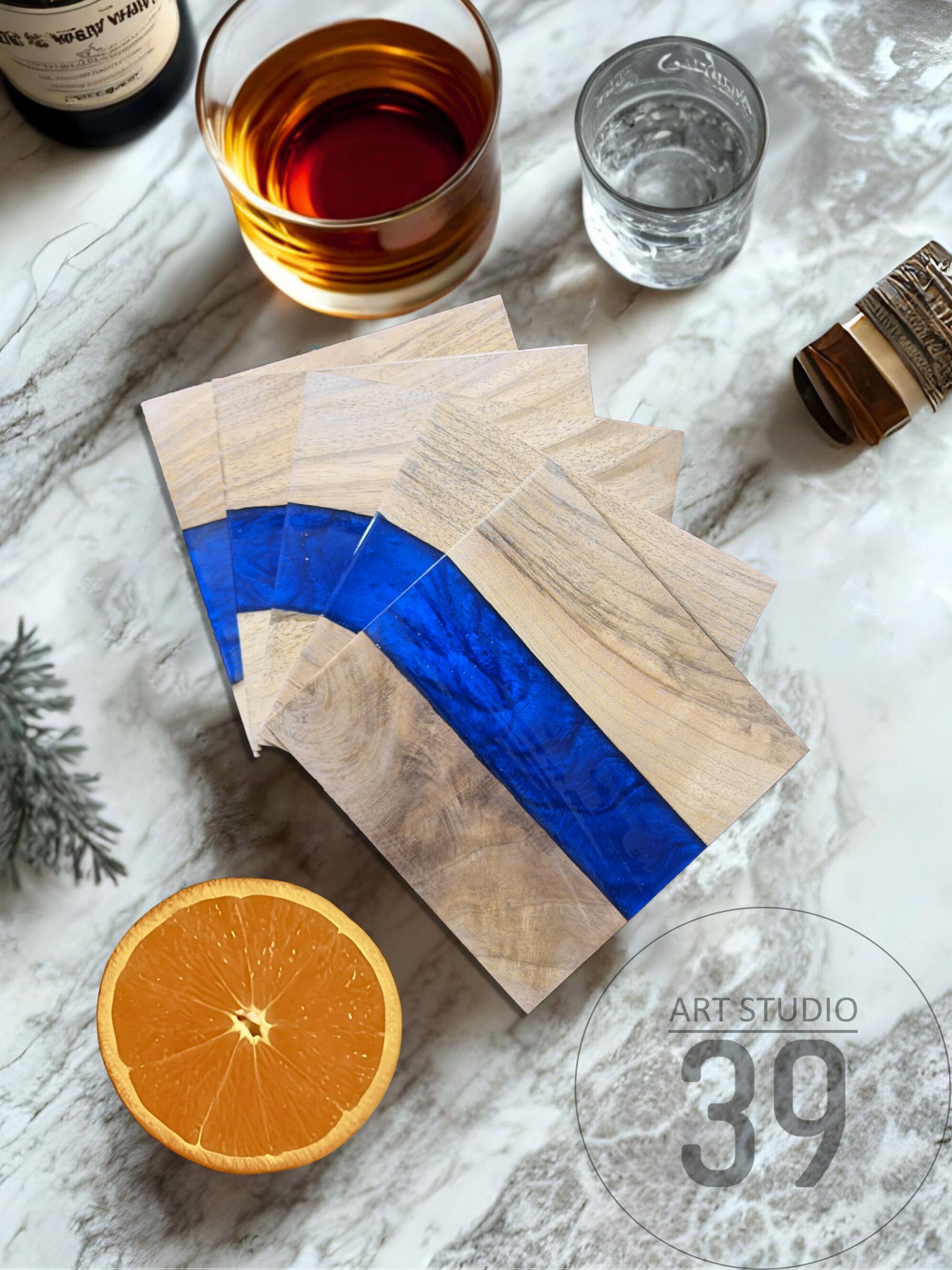Wooden coasters with a blue resin river design. The coasters are made of light-colored wood and feature a vibrant blue stripe. The image also includes a glass of amber liquid, a glass of water, and a sliced orange.