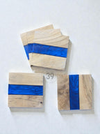 Wooden coasters with a blue resin stripe. The coasters are square-shaped and made of light-colored wood. The blue resin adds a pop of color and a modern touch. The coasters are arranged on a white surface.