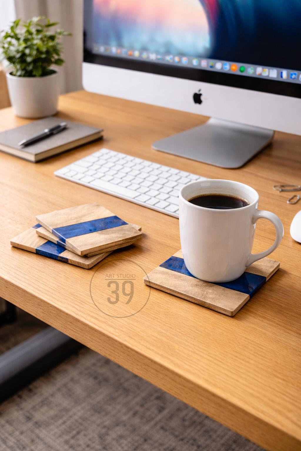 Wooden coasters with a blue stripe, paired with a white coffee mug filled with dark liquid. The coasters are on a wooden desk, near a computer and keyboard. The coasters have the text "ART STUDIO 39".
