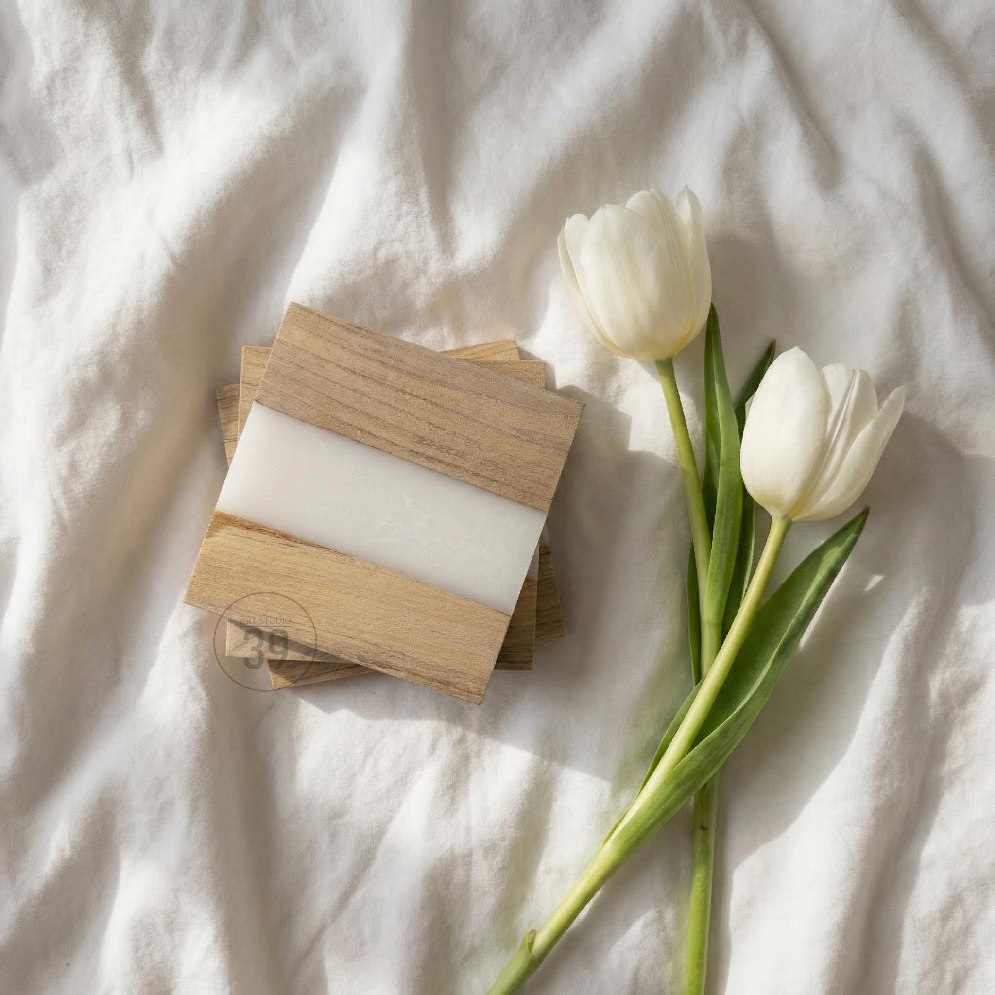 A stack of square coasters with a wood grain design and a white resin stripe. Two white tulips with green stems and leaves are next to the coasters. The coasters are on a white, textured surface.
