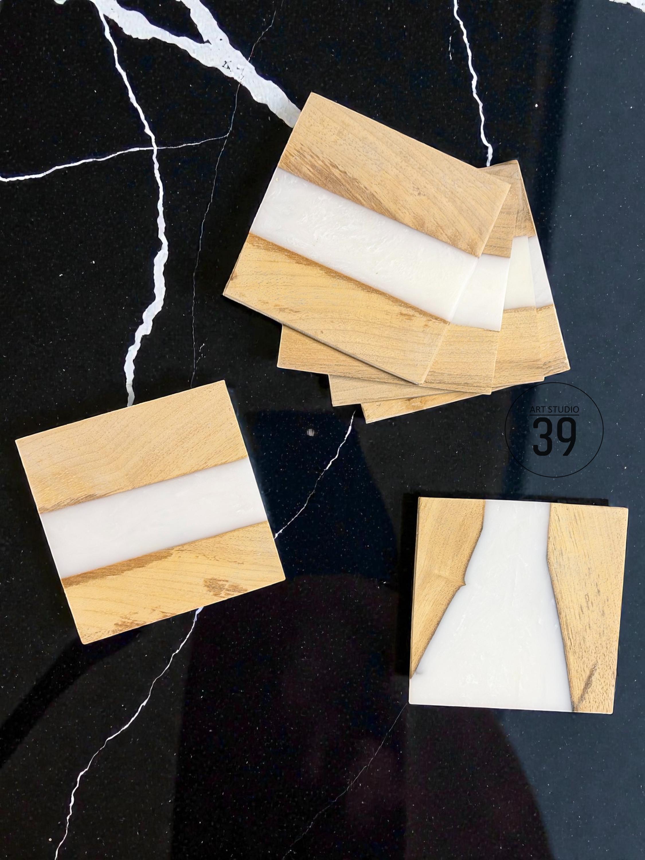 Square wooden coasters with a white resin inlay. The coasters have a natural wood grain finish and a smooth, glossy white center. The coasters are arranged on a black surface with white veining.