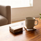 A stack of square wooden coasters with a white stripe and a beige mug on a wooden table. The coasters are made of wood and have a natural finish. The mug is beige and has a simple design. The text "ART STUDIO 39" is in a white circle.