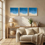 Three square wall art pieces depicting a beach scene with blue ocean and sandy shore. The artwork is displayed above a wooden console table with a lamp, books, and a woven basket. The text "ART STUDIO 39" is in the center.