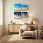 Four square wall art pieces depicting ocean scenes with blue, white, and gold resin. The artwork is displayed on a neutral wall above a wooden cabinet and a beige sofa. The room is decorated with a rug, lamp, and decorative items.