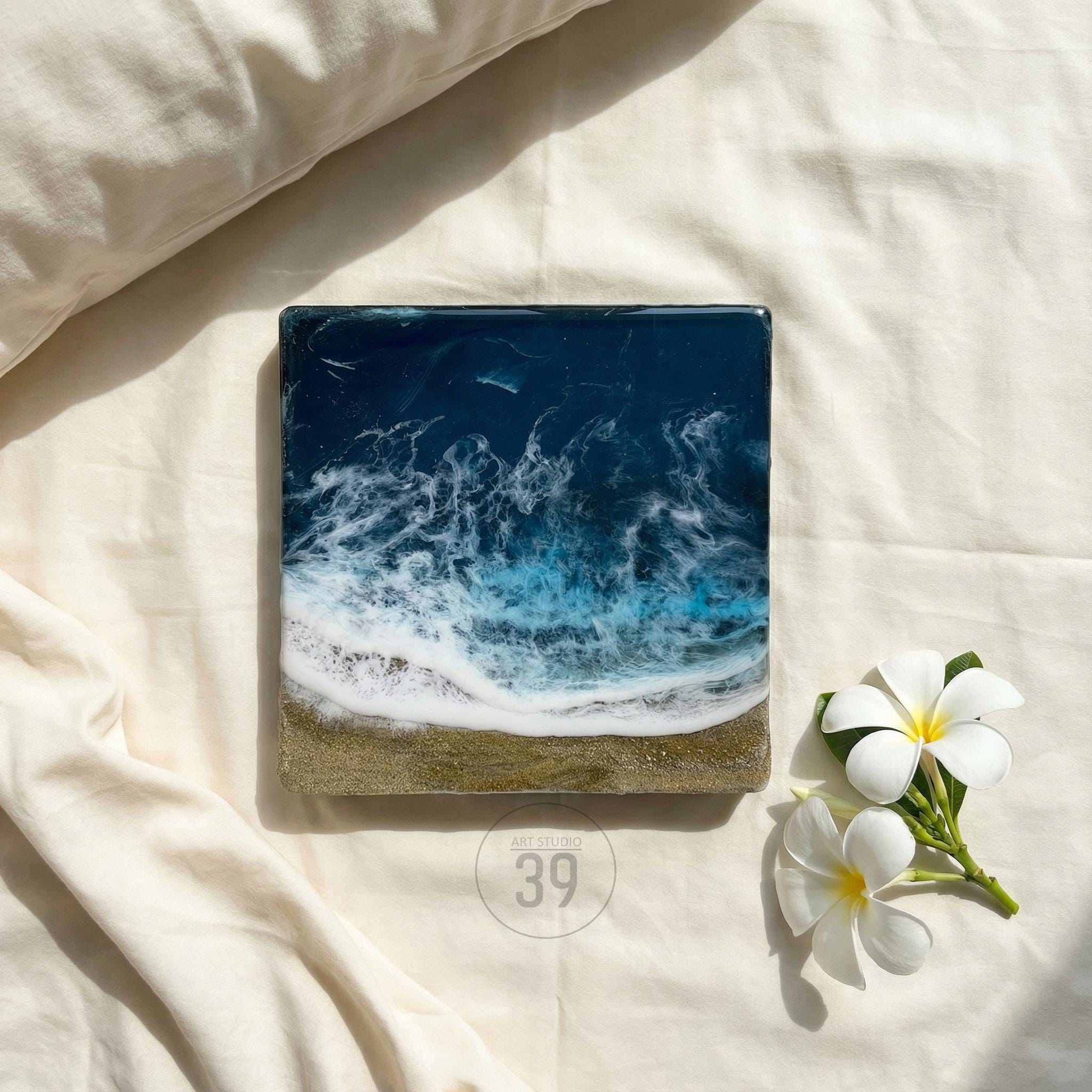 Square resin art piece depicting an ocean scene with foamy white waves crashing onto a sandy shore. The artwork features shades of blue, white, and gold. The number "39" is printed on the bottom. Two white flowers with yellow centers are in the lower right corner.