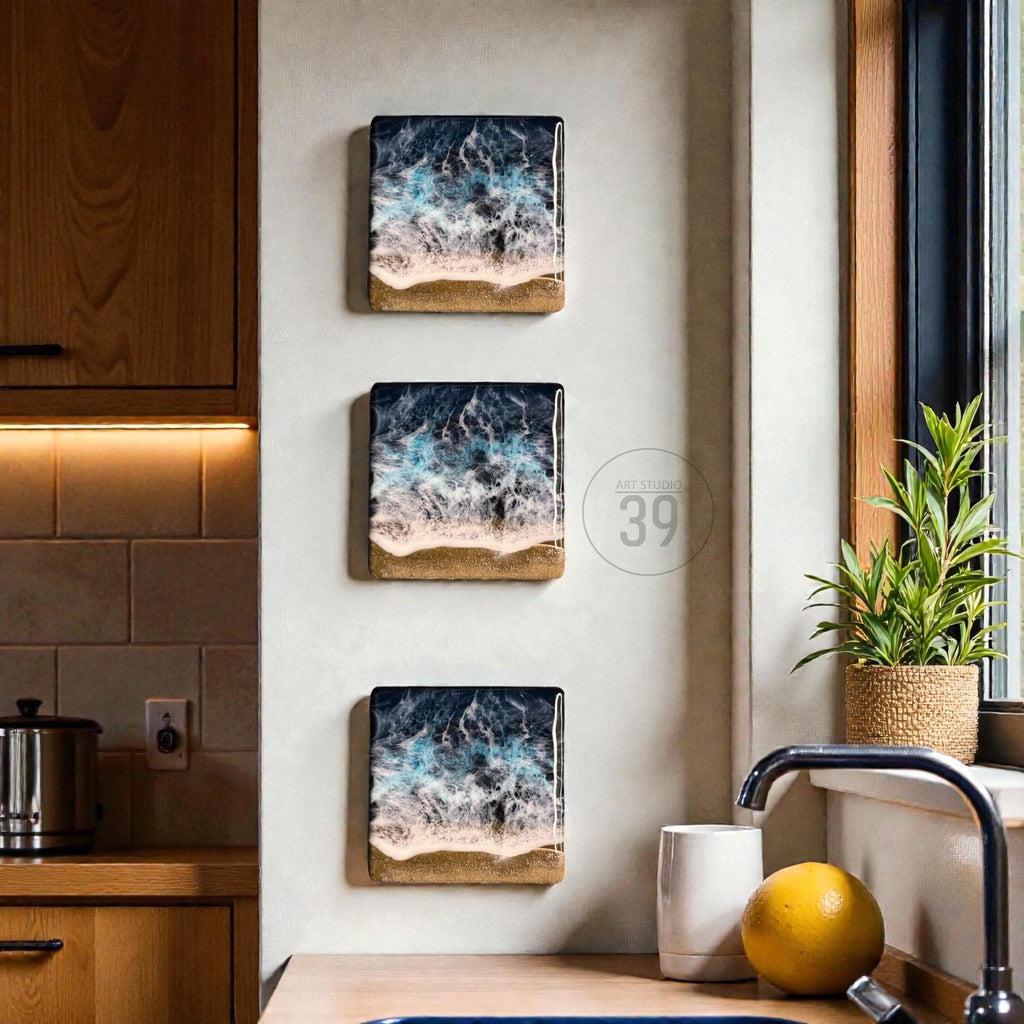 Three rectangular wall art pieces depicting an ocean scene with crashing waves and a sandy beach. The artwork features shades of blue, white, and tan, creating a coastal theme. The pieces are mounted on a light-colored wall.