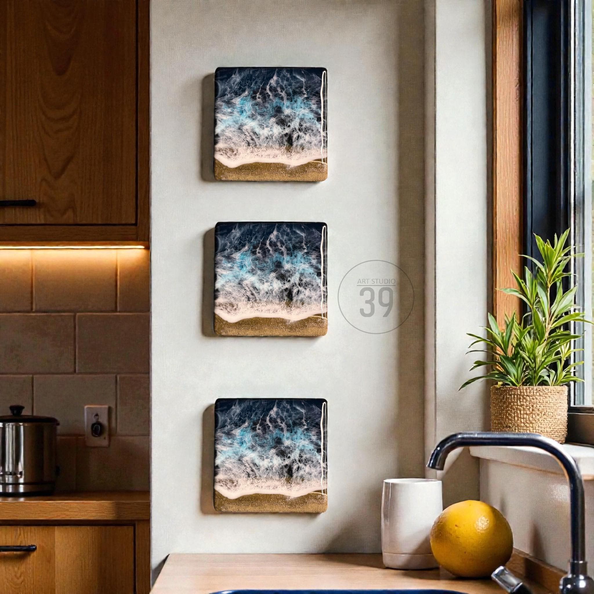 Three rectangular wall art pieces depicting an ocean scene with crashing waves and a sandy beach. The artwork features shades of blue, white, and tan, creating a coastal theme. The pieces are mounted on a light-colored wall.