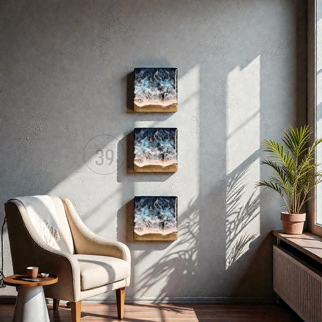 Three square wall art pieces depicting ocean waves with blue, white, and tan hues. The artwork is mounted on a gray wall, with a beige armchair and a small side table in the foreground. A potted plant sits near a window.