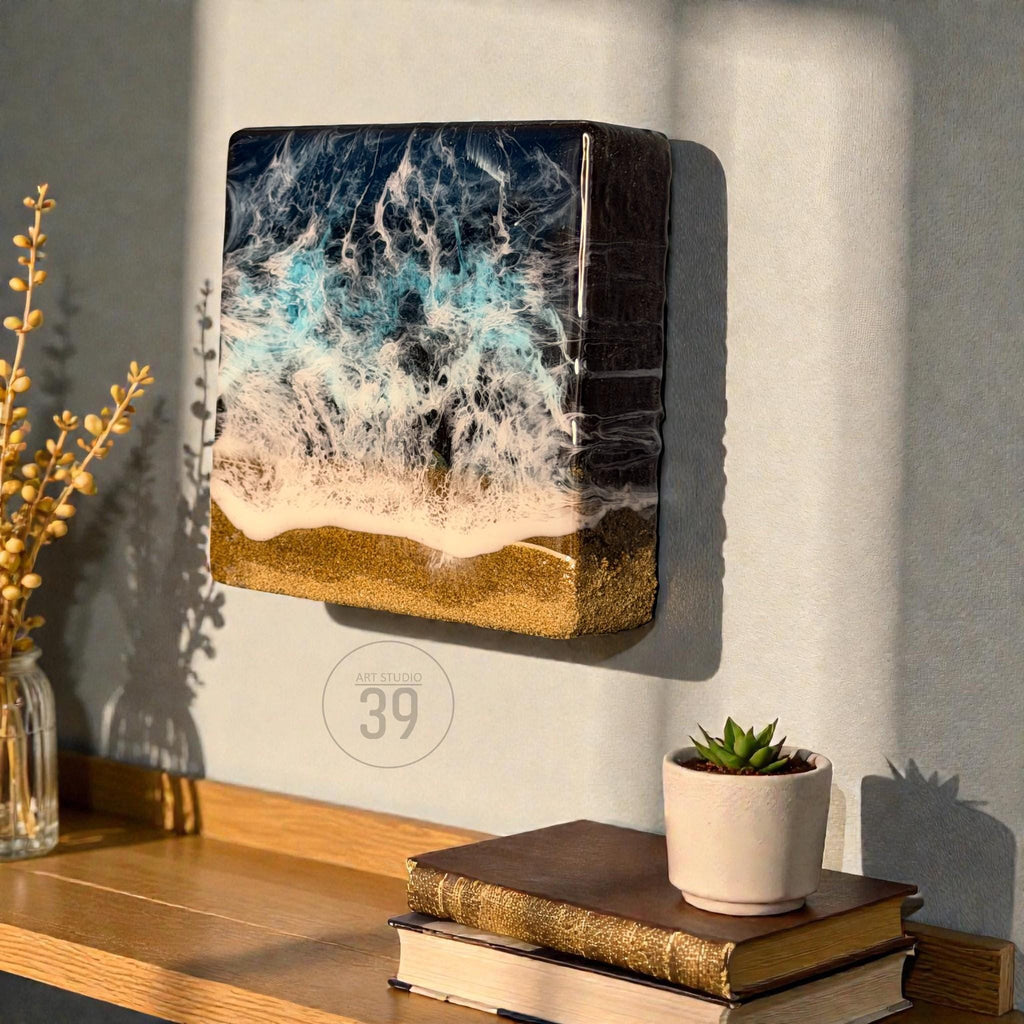 A rectangular wall art piece depicting an ocean scene. The artwork features a dark blue and turquoise wave design with white foam, transitioning to a golden sand base. The piece is mounted on a light gray wall.