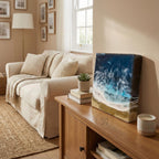 A rectangular resin art piece depicting an ocean scene with white waves, blue water, and a sandy shore. The artwork is displayed on a wooden cabinet next to a lit candle and a small succulent plant.