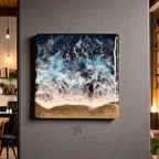 Square resin art piece depicting an ocean scene. The artwork features a dark blue and turquoise wave design with white foam, transitioning to a golden sand base. The piece is mounted on a gray wall.