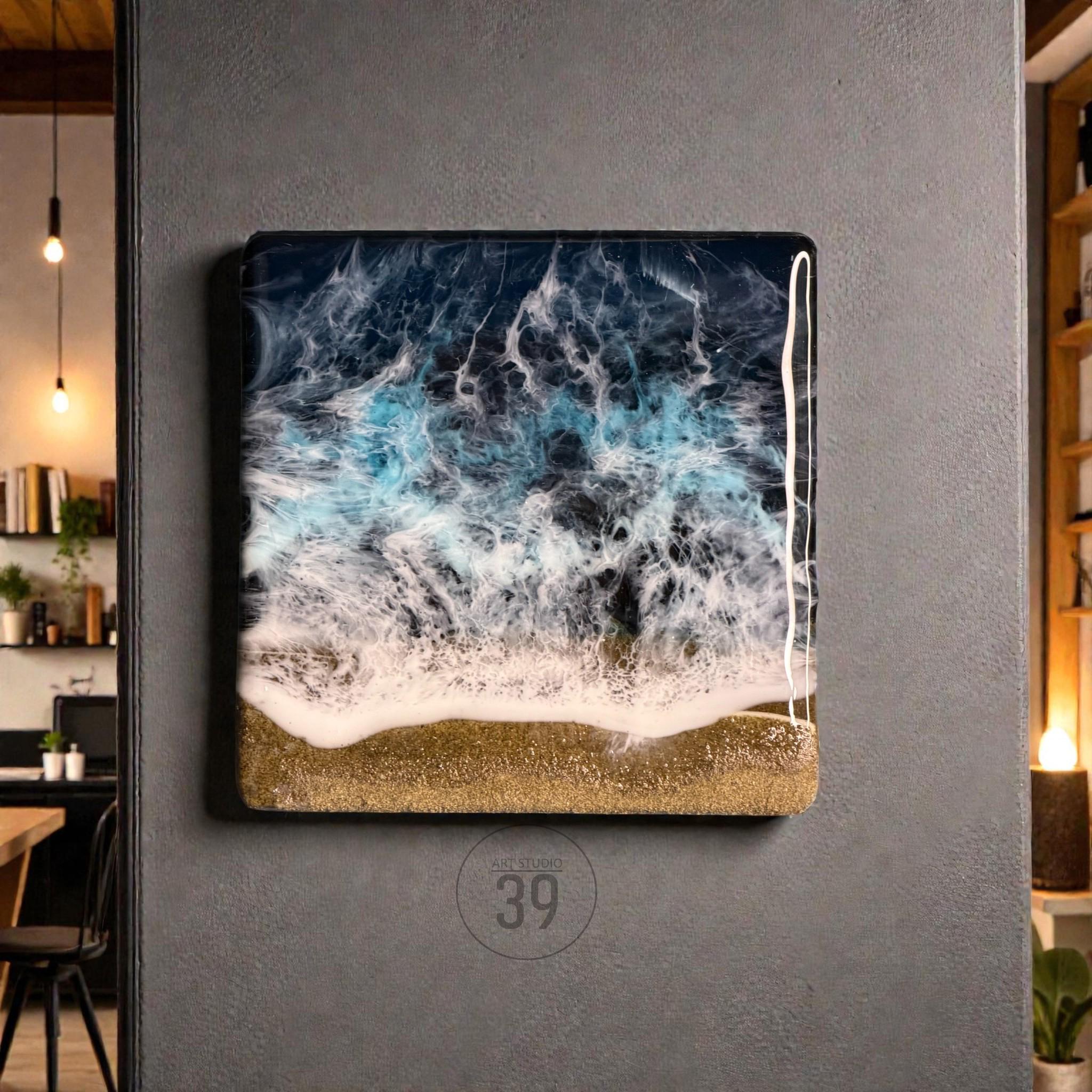 Square resin art piece depicting an ocean scene. The artwork features a dark blue and turquoise wave design with white foam, transitioning to a golden sand base. The piece is mounted on a gray wall.