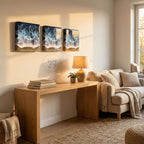 Three framed wall art pieces depicting ocean waves with blue, white, and tan colors. A wooden desk with books, a lamp, and a plant are in front of a beige sofa. The wall art is above the desk.