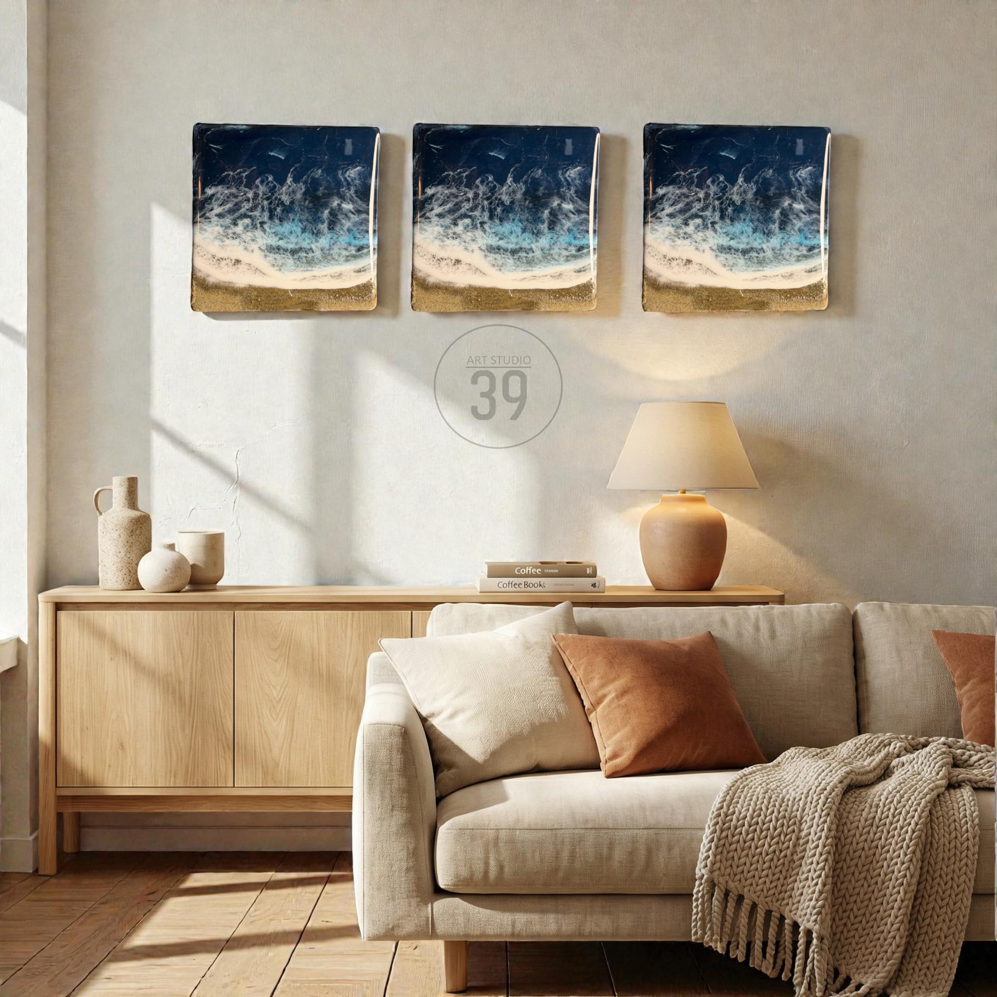 Three square wall art pieces depicting an ocean scene with dark blue water, white waves, and a golden sand beach. The art is displayed on a light-colored wall above a wooden cabinet and a beige sofa. The text "Art studio 39" is in the center.