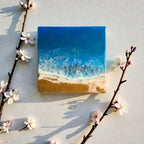 Square resin art piece depicting a beach scene with a blue ocean, white foamy waves, and a sandy shore. The artwork is surrounded by white flowers and branches. The text "39" is visible.