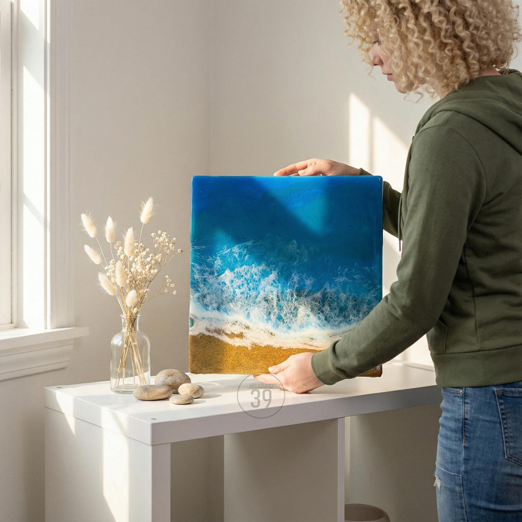 Square resin art piece depicting an aerial view of a beach scene. The artwork features a vibrant blue ocean with white foamy waves crashing onto a golden sand beach. The piece is held by a person, showcasing the artwork's size and detail.