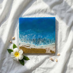 Square resin art piece depicting an ocean scene with blue waves, a sandy shore, and white foam. A white flower with a yellow center and seashells are arranged around the artwork. The text "ART STUDIO 39" is visible.