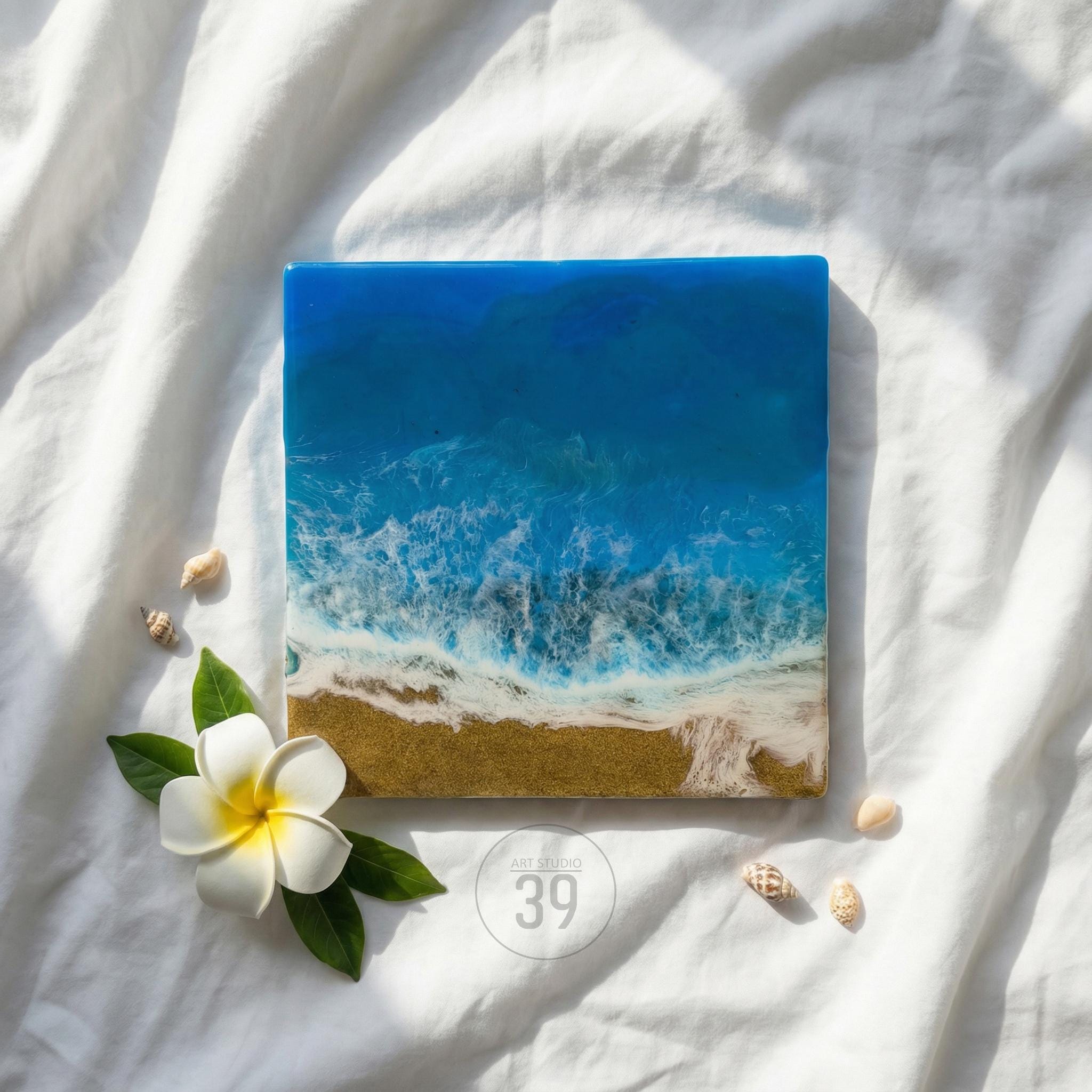 Square resin art piece depicting an ocean scene with blue waves, a sandy shore, and white foam. A white flower with a yellow center and seashells are arranged around the artwork. The text "ART STUDIO 39" is visible.