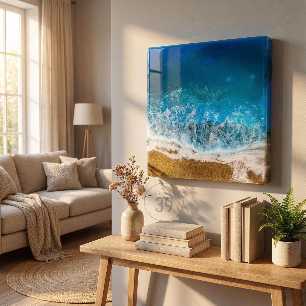 A square wall art piece depicting a beach scene with blue ocean waves, white foam, and golden sand. The artwork is framed and mounted on a neutral-colored wall. The room also features a beige sofa, wooden table, and decorative items.