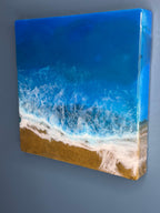 Square resin art piece depicting a beach scene. The artwork features a gradient of blues representing the ocean, white foamy waves, and a golden sand beach. The piece is mounted on a square frame.