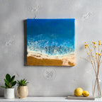 Square resin art piece depicting a beach scene. The artwork features a deep blue ocean, white foamy waves, and a golden sand beach. The piece is mounted on a wall, with plants and lemons in the foreground.