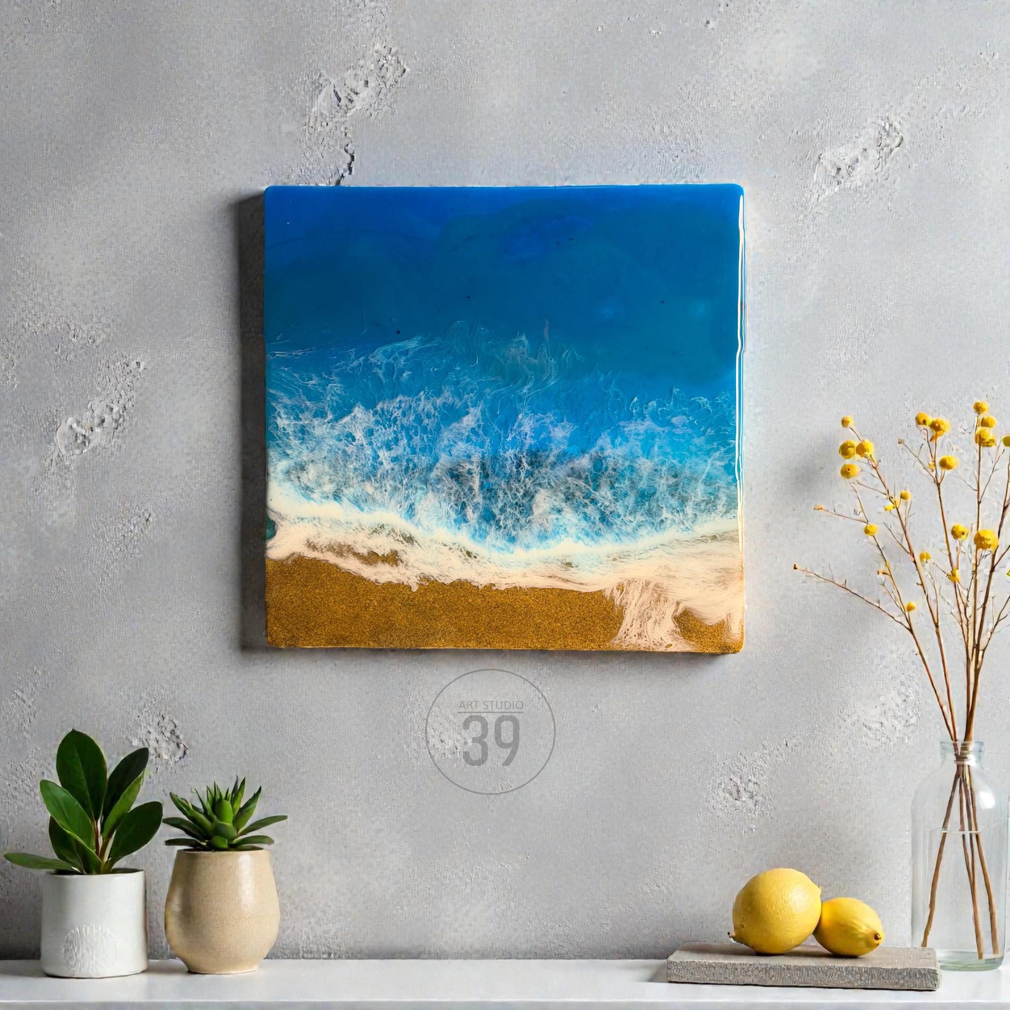 Square resin art piece depicting a beach scene. The artwork features a deep blue ocean, white foamy waves, and a golden sand beach. The piece is mounted on a wall, with plants and lemons in the foreground.