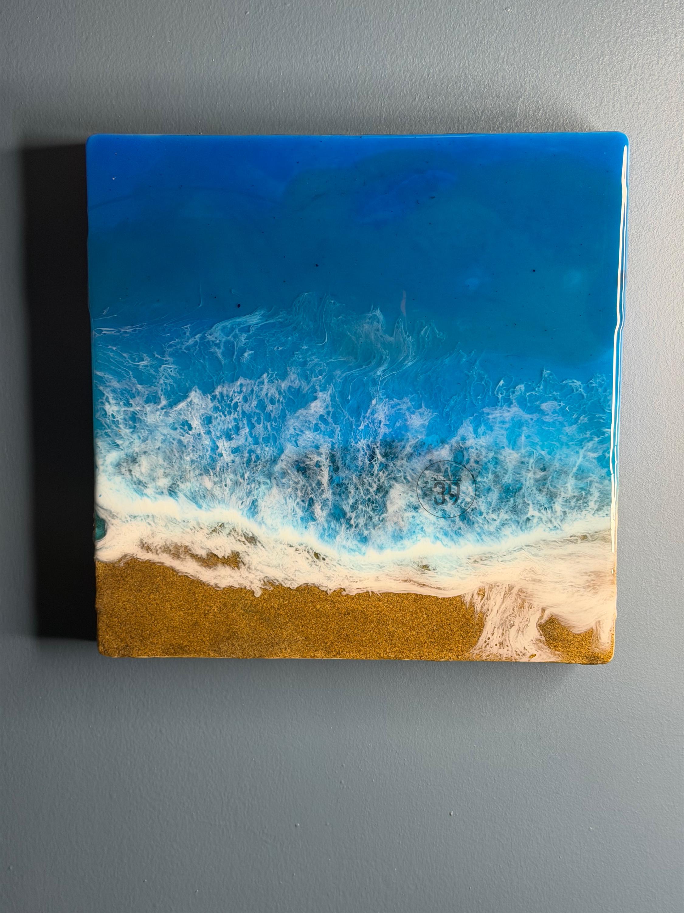 Square resin art piece depicting a beach scene. The top portion is a deep blue, transitioning to lighter shades, representing the ocean. White foamy waves crash onto a golden sand beach at the bottom. The artwork is mounted on a gray wall.