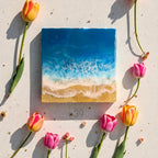 Square resin art piece depicting a beach scene with blue ocean waves and golden sand. The artwork is surrounded by pink and yellow tulips. The piece has the text "ART STUDIO 39" in a circle.