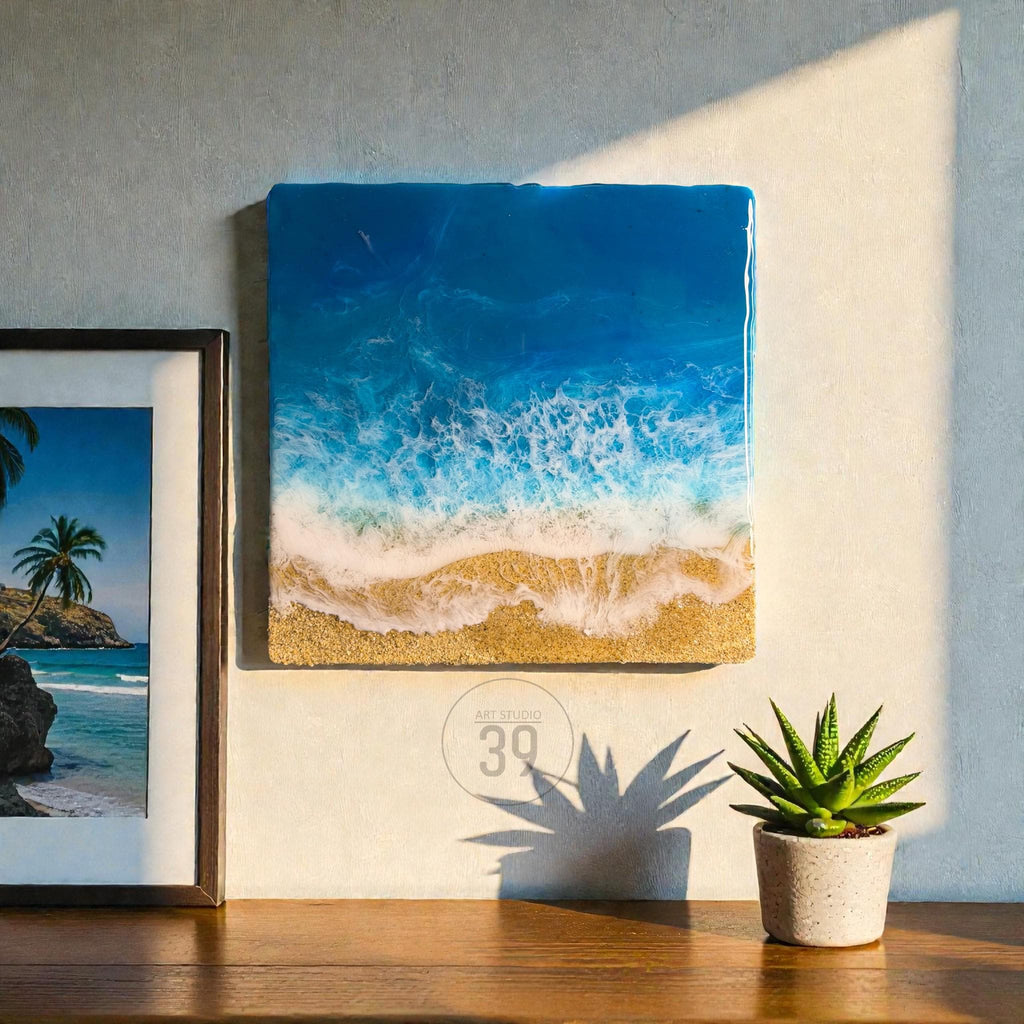 Square resin art piece depicting an ocean scene with blue and white waves crashing onto a sandy beach. The artwork is mounted on a wall, next to a framed picture of a tropical beach. A small potted succulent sits on a wooden surface.