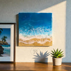 Square resin art piece depicting an ocean scene with blue and white waves crashing onto a sandy beach. The artwork is mounted on a wall, next to a framed picture of a tropical beach. A small potted succulent sits on a wooden surface.