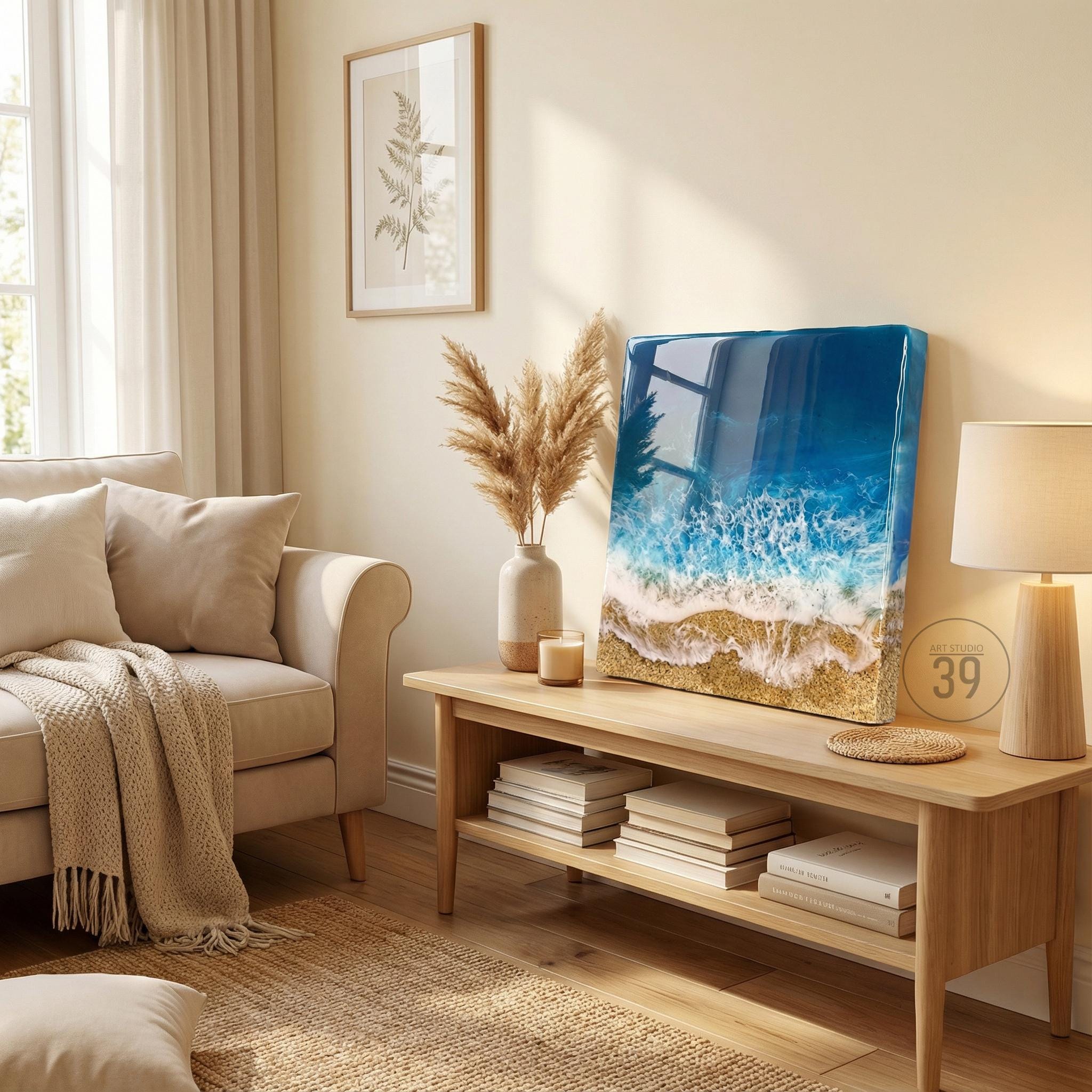 A square resin art piece depicting a beach scene with blue ocean waves, white foam, and golden sand. The artwork is displayed on a light wood table with books and a lamp. A beige sofa and framed botanical print are in the background.