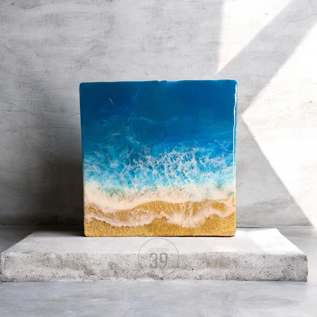 Square resin art piece depicting an ocean scene. The top is a deep blue, transitioning to white foamy waves, and ending with a sandy beach. The artwork is displayed on a concrete surface.