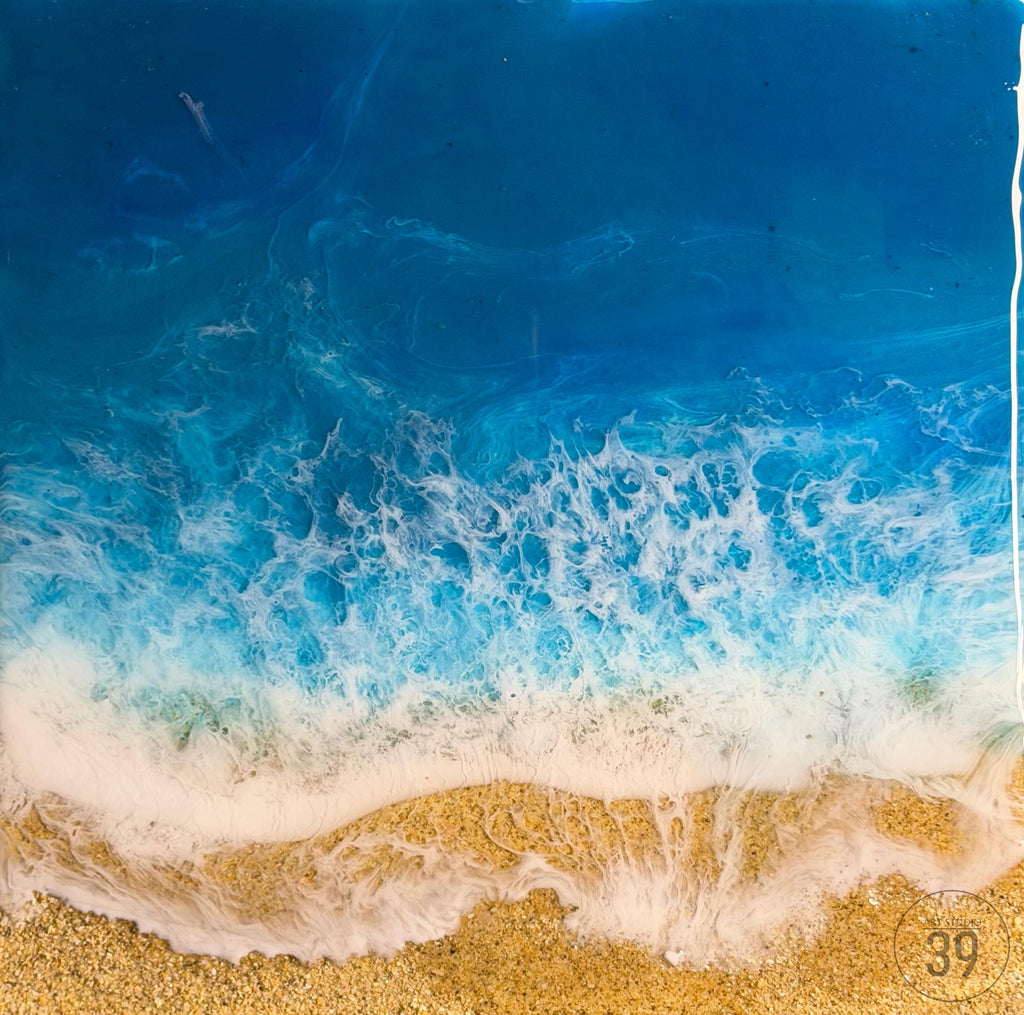 Abstract resin art depicting a beach scene. The artwork features a gradient of deep blue to turquoise, representing the ocean, with white foamy waves crashing onto a sandy shore. The sand is a textured golden color.