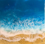 Abstract resin art depicting a beach scene. The artwork features a gradient of deep blue to turquoise, representing the ocean, with white foamy waves crashing onto a sandy shore. The sand is a textured golden color.