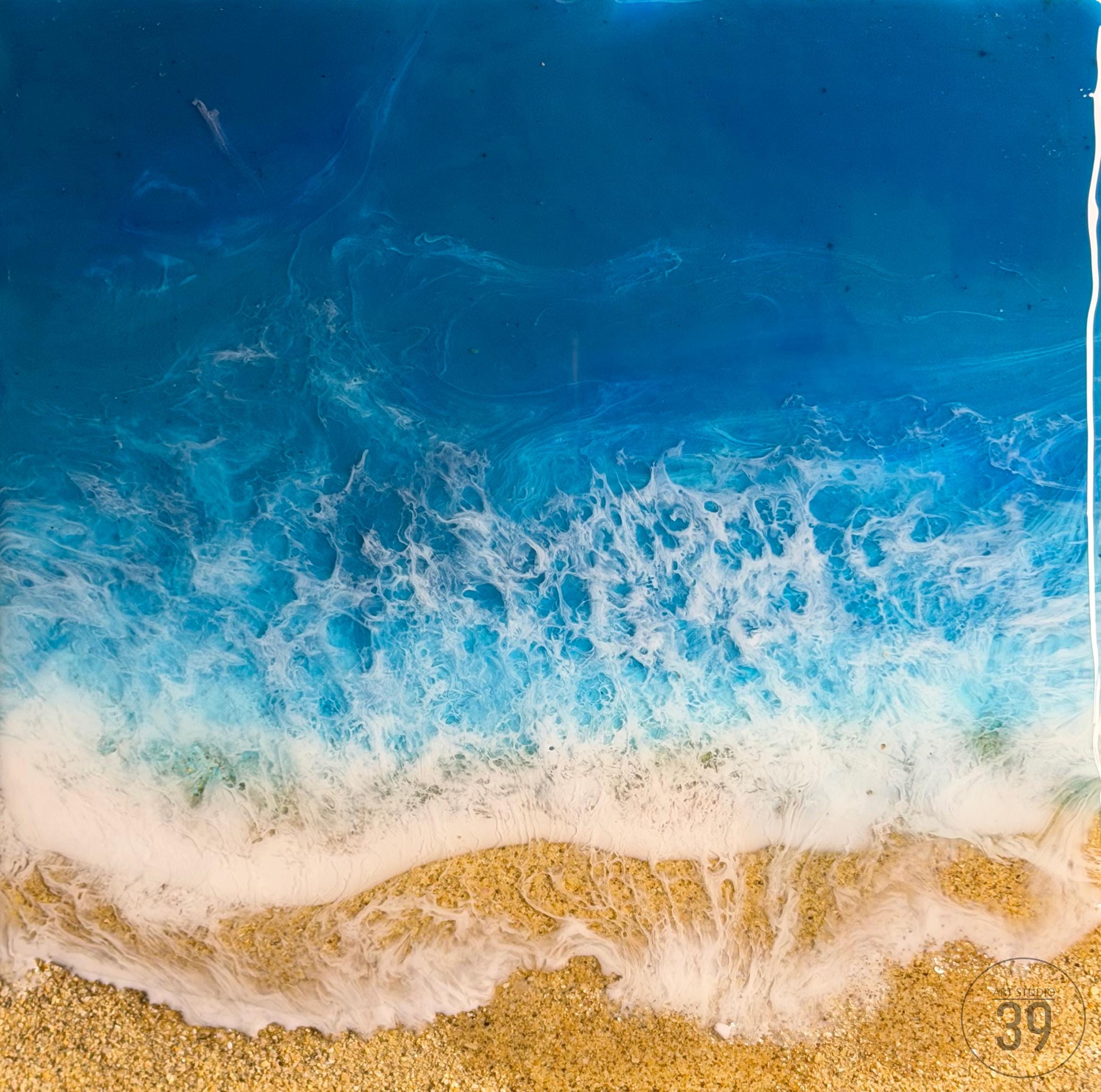 Abstract resin art depicting a beach scene. The artwork features a gradient of deep blue to turquoise, representing the ocean, with white foamy waves crashing onto a sandy shore. The sand is a textured golden color.