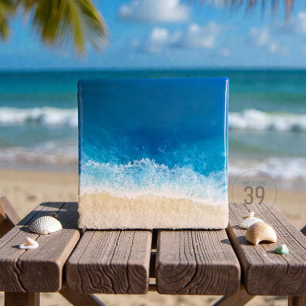 Square resin art piece depicting an ocean scene with a sandy beach and foamy waves. The artwork features shades of blue and white, with a sandy-colored base. The piece is displayed on a wooden table with seashells.