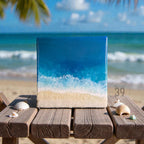 Square resin art piece depicting an ocean scene with a sandy beach and foamy waves. The artwork features shades of blue and white, with a sandy-colored base. The piece is displayed on a wooden table with seashells.