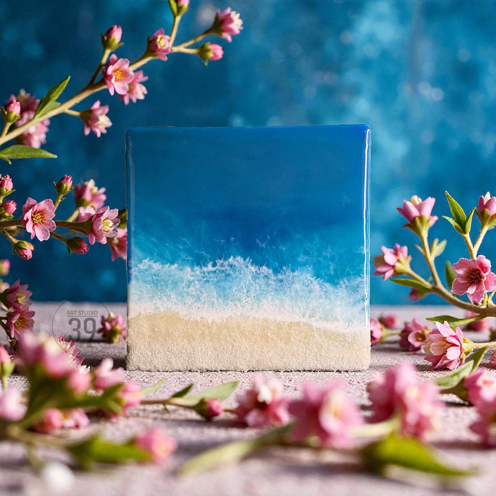 Square resin art piece depicting a beach scene. The artwork features a gradient of blues representing the ocean, white foamy waves, and a sandy beach. Pink flowers and branches frame the artwork.