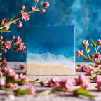 Square resin art piece depicting a beach scene. The artwork features a gradient of blues representing the ocean, white foamy waves, and a sandy beach. Pink flowers and branches frame the artwork.
