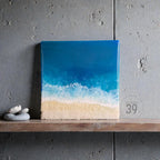 Square artwork depicting a beach scene with a gradient of blue ocean and white foamy waves meeting a sandy shore. The artwork is displayed on a wooden shelf against a gray wall. The text "ART STUDIO 39." is in the lower right corner.