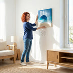 A person hanging a framed abstract art piece. The artwork features a blue and white ocean scene with a sandy beach at the bottom. The frame is a light color, and the artwork is being hung on a white wall. The text "ART STUDIO 39" is visible.