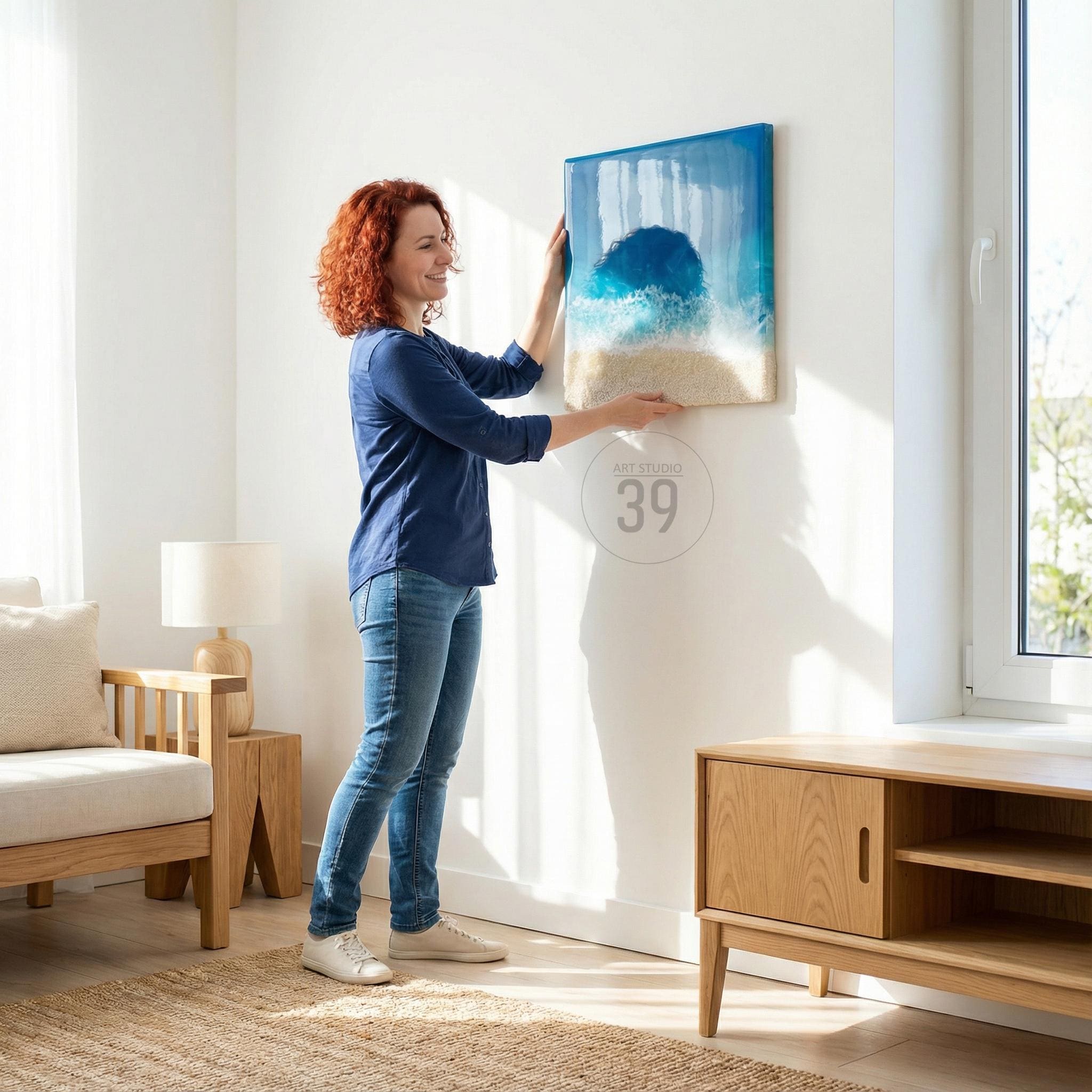 A person hanging a framed abstract art piece. The artwork features a blue and white ocean scene with a sandy beach at the bottom. The frame is a light color, and the artwork is being hung on a white wall. The text "ART STUDIO 39" is visible.
