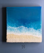 Square resin art piece depicting a beach scene. The top portion is a gradient of blues representing the ocean, transitioning to white foamy waves, and a sandy textured beach at the bottom. The text "ART STUDIO 39" is in a circle at the bottom.