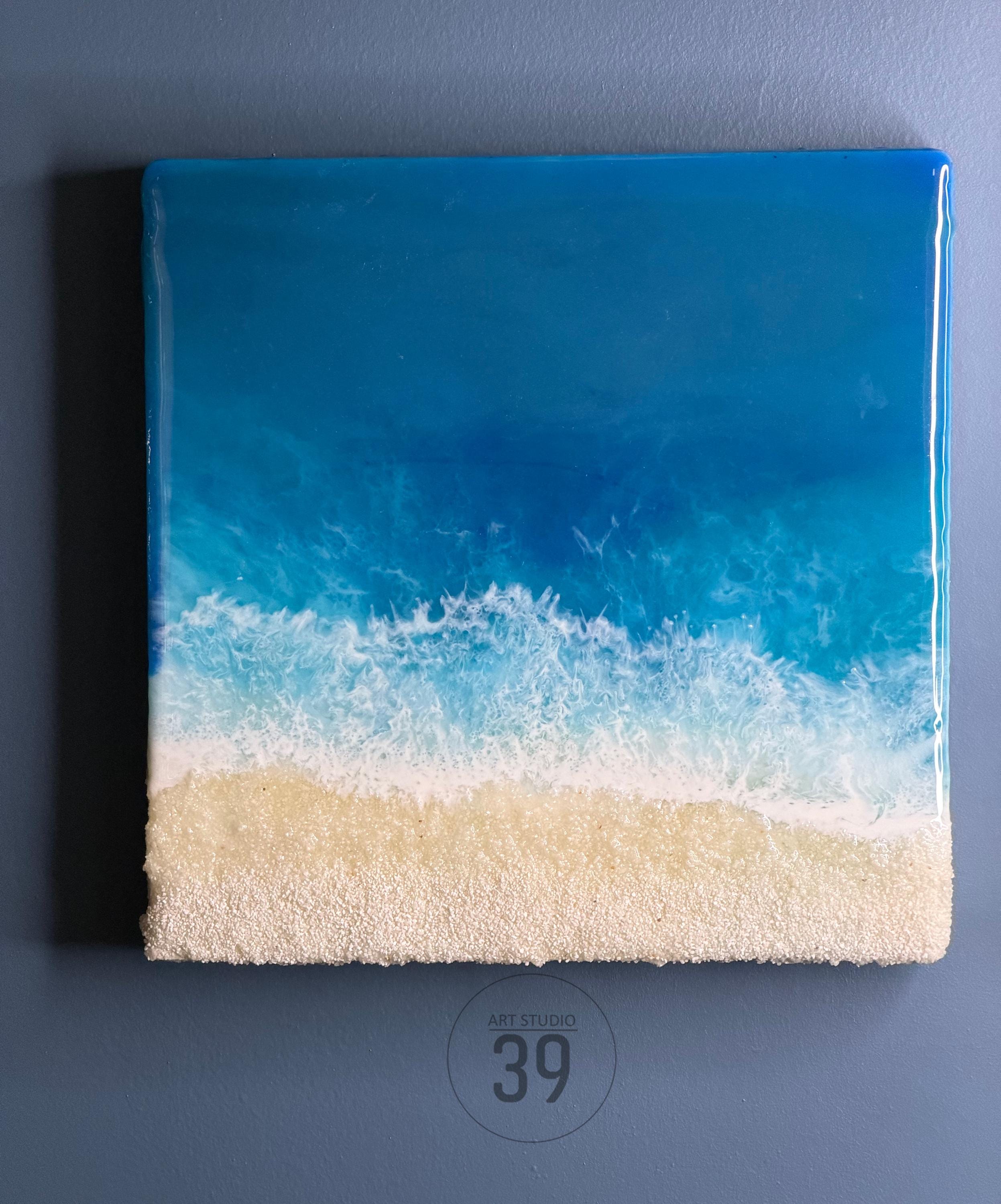 Square resin art piece depicting a beach scene. The top portion is a gradient of blues representing the ocean, transitioning to white foamy waves, and a sandy textured beach at the bottom. The text "ART STUDIO 39" is in a circle at the bottom.