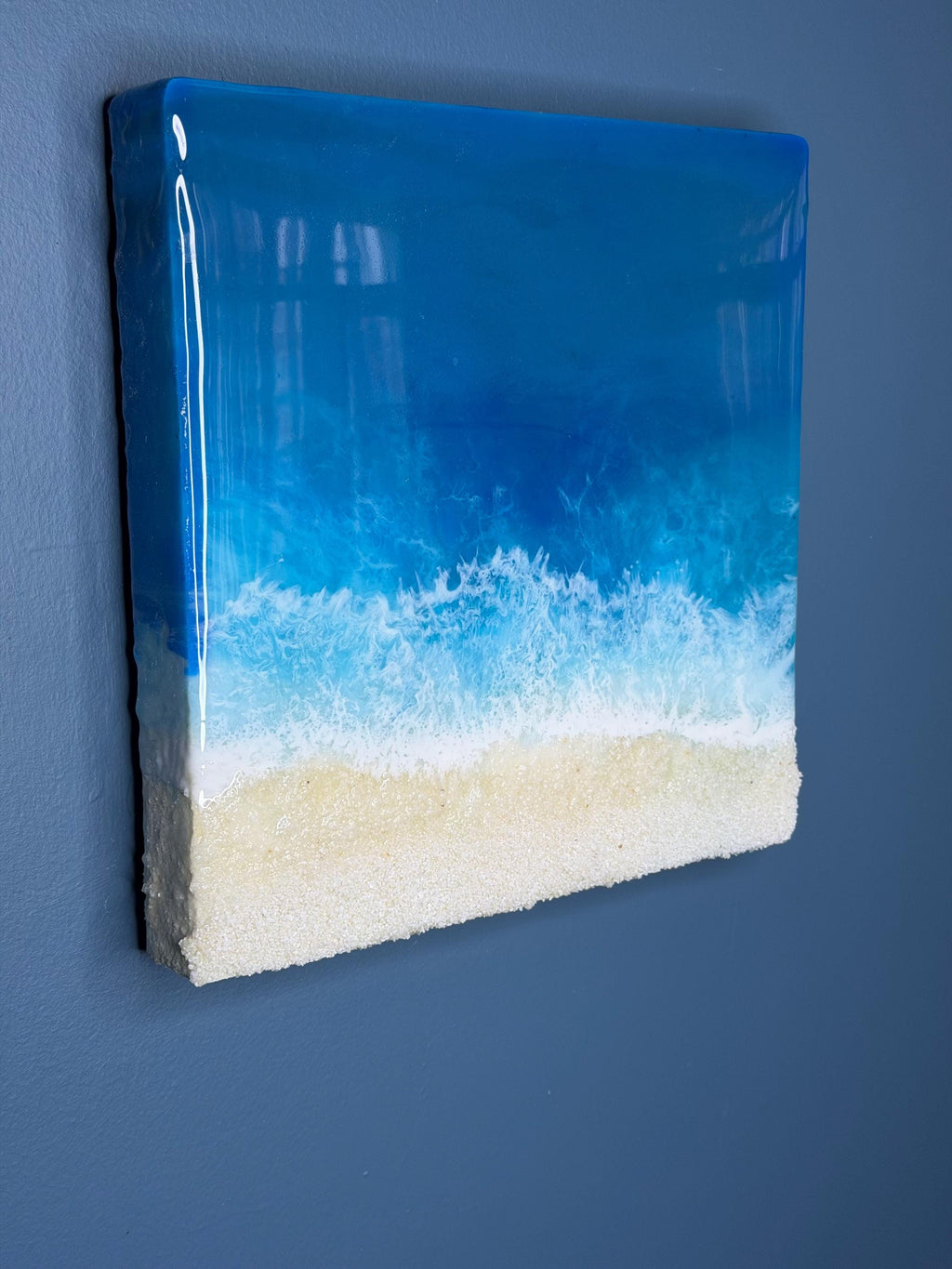 A square resin art piece depicting a beach scene. The top is a glossy, deep blue representing the ocean, transitioning to white foamy waves and a sandy beige beach at the bottom. The artwork is mounted on a white, textured base.
