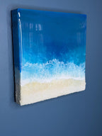 A square resin art piece depicting a beach scene. The top is a glossy, deep blue representing the ocean, transitioning to white foamy waves and a sandy beige beach at the bottom. The artwork is mounted on a white, textured base.