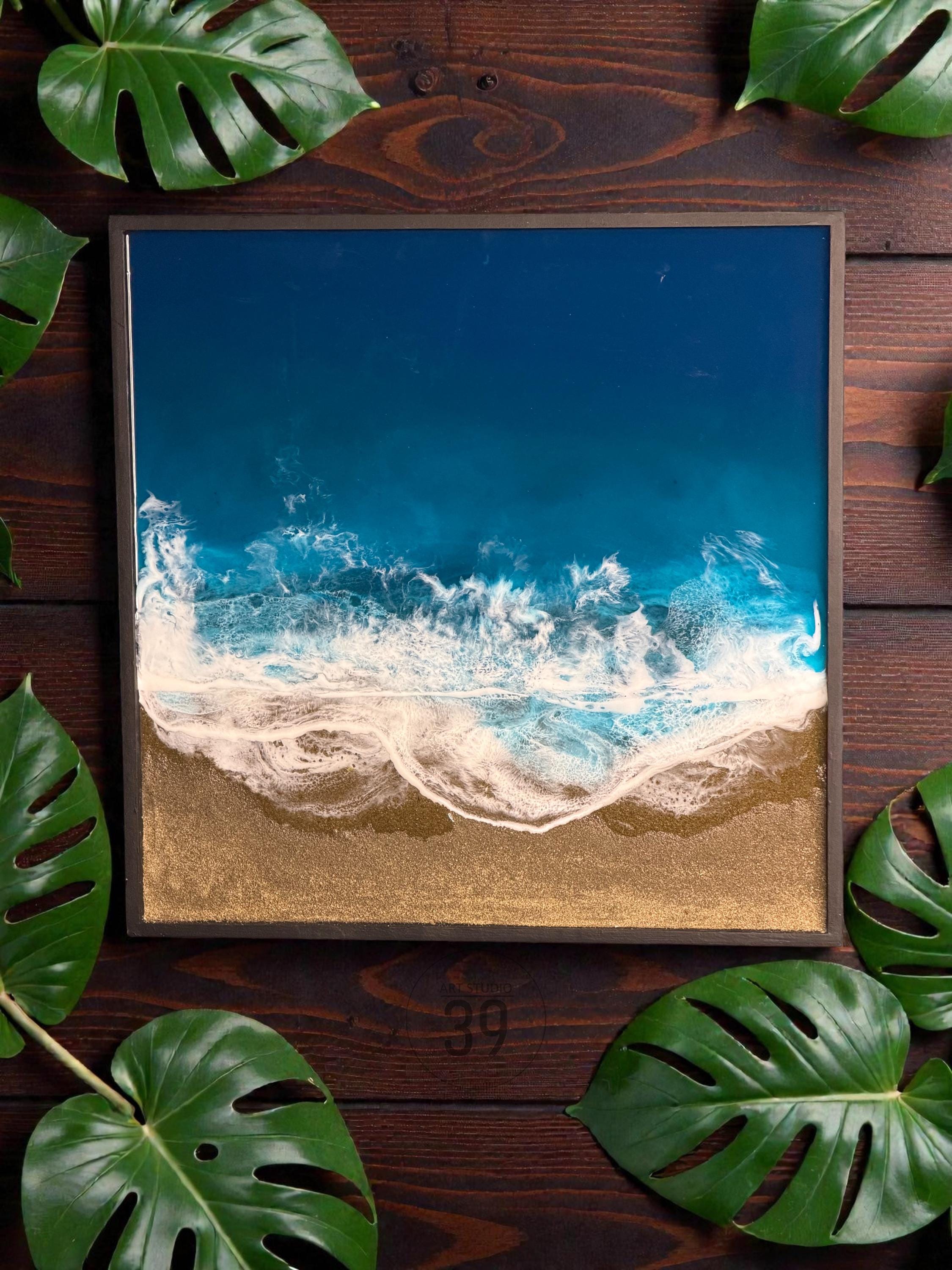 Square resin art piece depicting an aerial view of a beach scene. The artwork features a deep blue ocean, white foamy waves, and a golden sand beach. The piece is framed in a dark brown wooden frame.