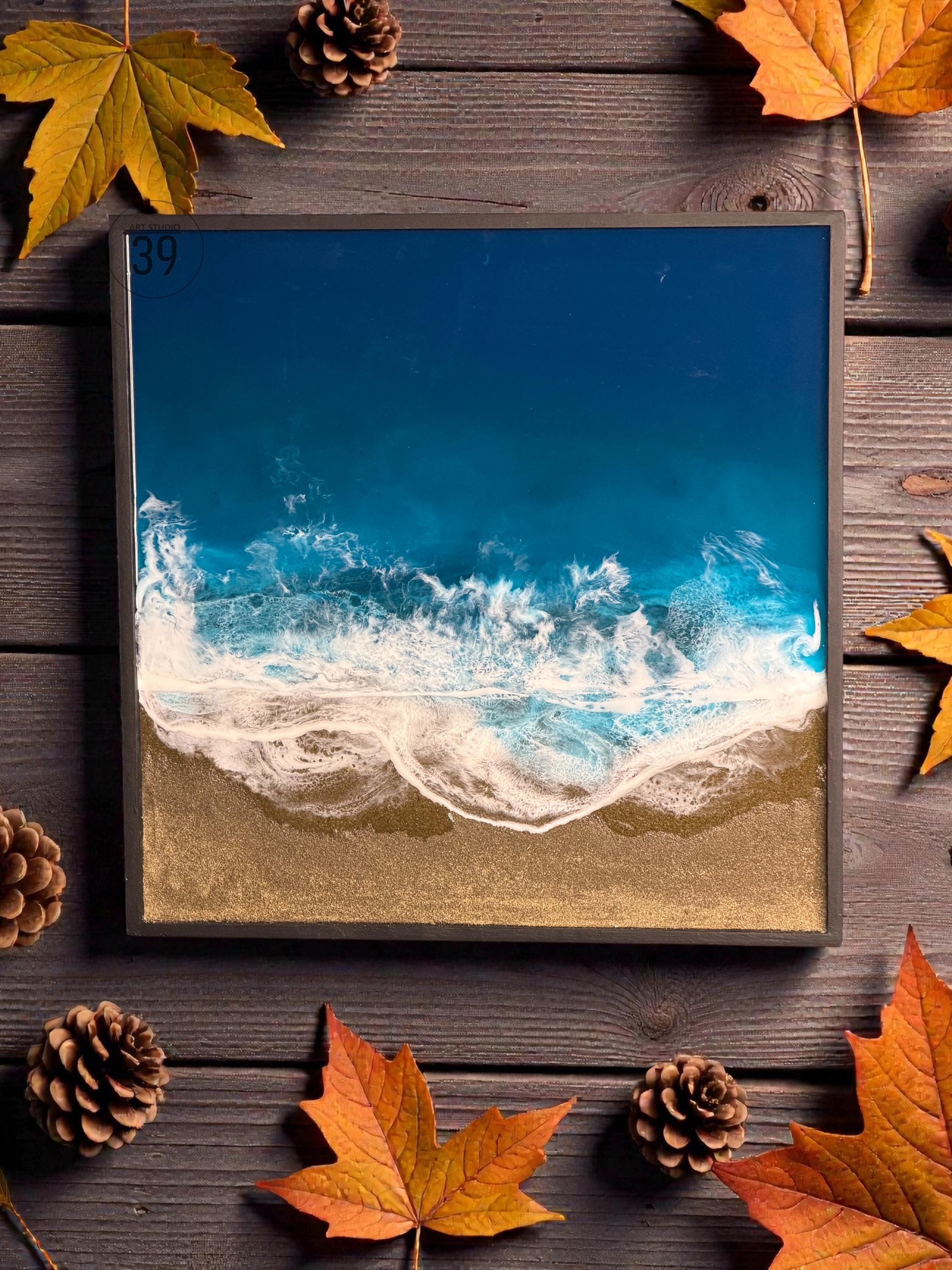 Square resin art piece depicting an ocean scene. The artwork features a deep blue sky transitioning to turquoise waves with white foam, meeting a golden sand beach. The piece is framed in a dark gray border.