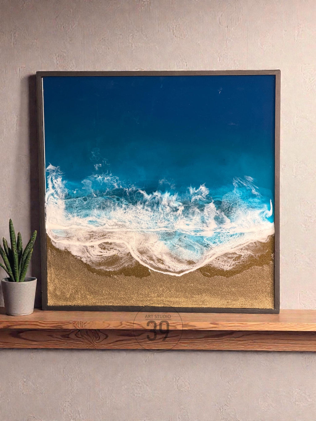 Square framed artwork depicting an aerial view of a beach scene. The artwork features a deep blue ocean, white foamy waves, and a golden sand shoreline. The piece is framed in a dark gray border and rests on a wooden shelf.