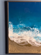 Ocean-themed artwork featuring a vibrant depiction of a beach scene. The artwork showcases a deep blue ocean with foamy white waves crashing onto a golden sandy shore. The piece is framed in a dark brown wooden frame.